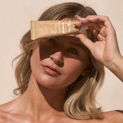 Woman holding a ThriveTallow tube of sunscreen near her face against a beige background