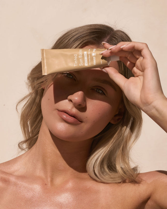 Woman holding a ThriveTallow tube of sunscreen near her face against a beige background