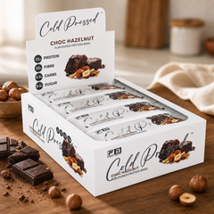 Fibre Boost Cold Pressed Choc Hazelnut Protein Bar box on a warm countertop with chocolate and hazelnuts